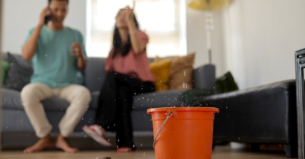A living room scene with an orange bucket collecting water from a ceiling leak, while two distressed individuals sit on a sofa in the background.
