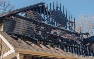 Close-up of a multi-story residential house with severe fire damage, including charred siding, exposed framing, and a partially collapsed roof under a clear blue sky.