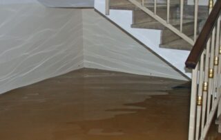Muddy brown floodwater fills a basement or ground-floor interior, partially submerging the base of a light stone staircase with a dark wood handrail and cream-colored walls.